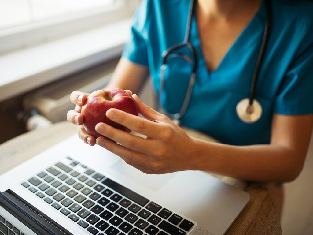 Nurse holding an apple while sitting at a desk with a laptop and a stethoscope